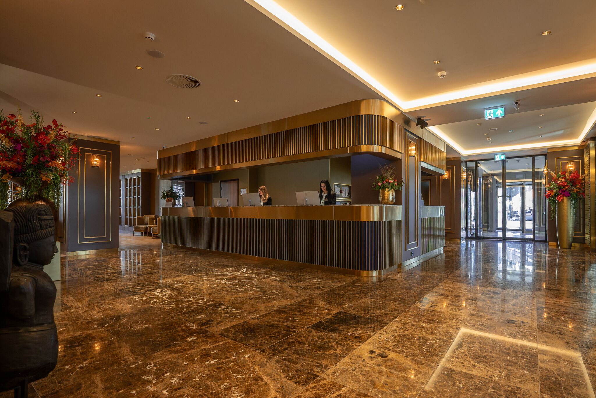 Een moderne hotellobby met een receptiebalie, marmeren vloeren en decoratieve planten.