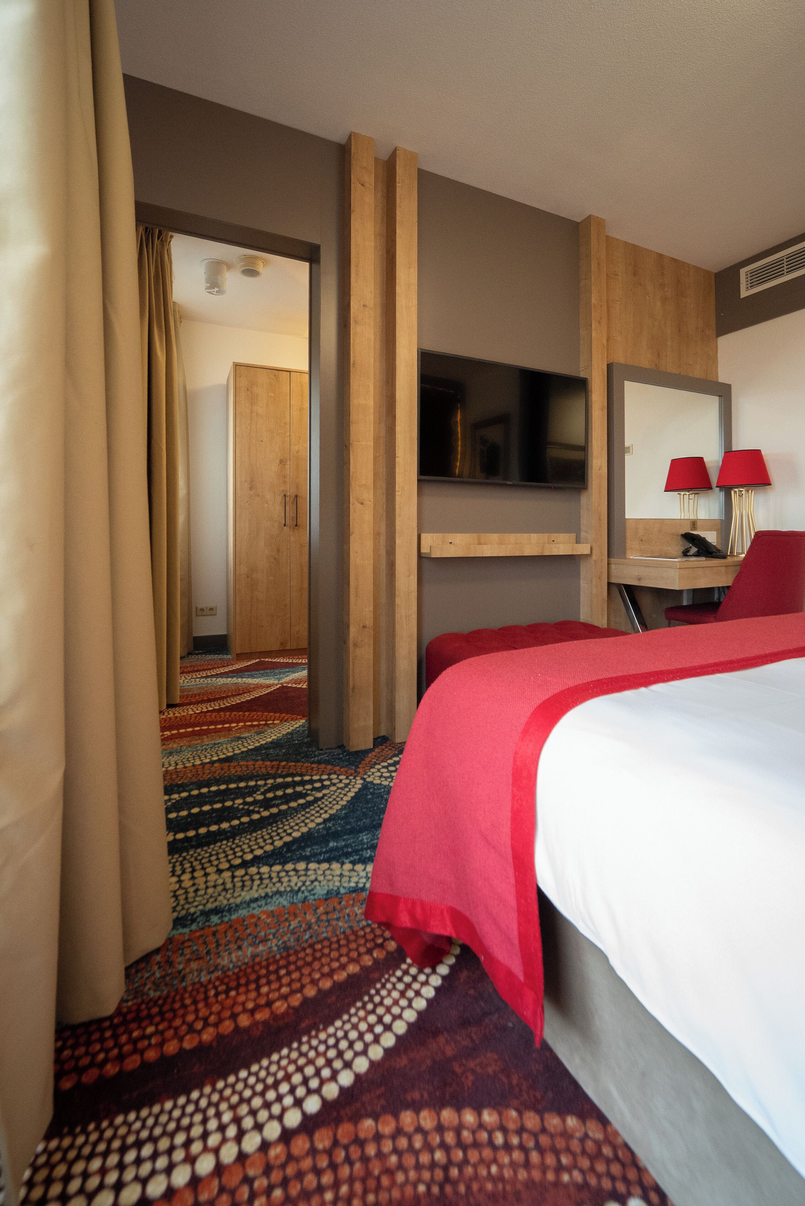 A hotel room with a large bed, a desk with a chair, a television, and a mirror. There is a door leading to another room and a patterned carpet.