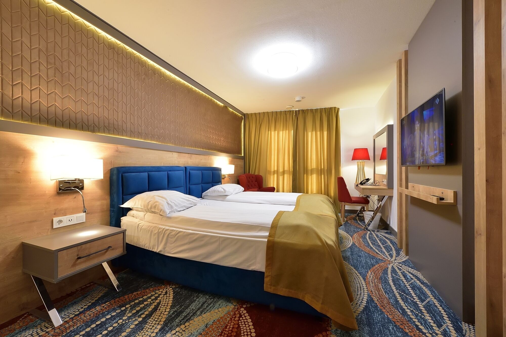Une chambre d’hôtel avec un grand lit, une table de chevet, un bureau avec une chaise, une télévision et une moquette à motifs.