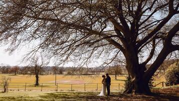 Una pareja está de pie bajo un árbol grande en un campo, con una valla al fondo.