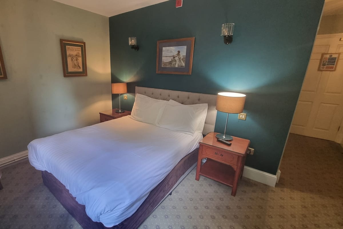 Ein ordentlich bezogenes Bett in einem Hotelzimmer, mit Kopfteil, zwei Nachttischen mit Lampen und zwei gerahmten Bildern an der Wand.