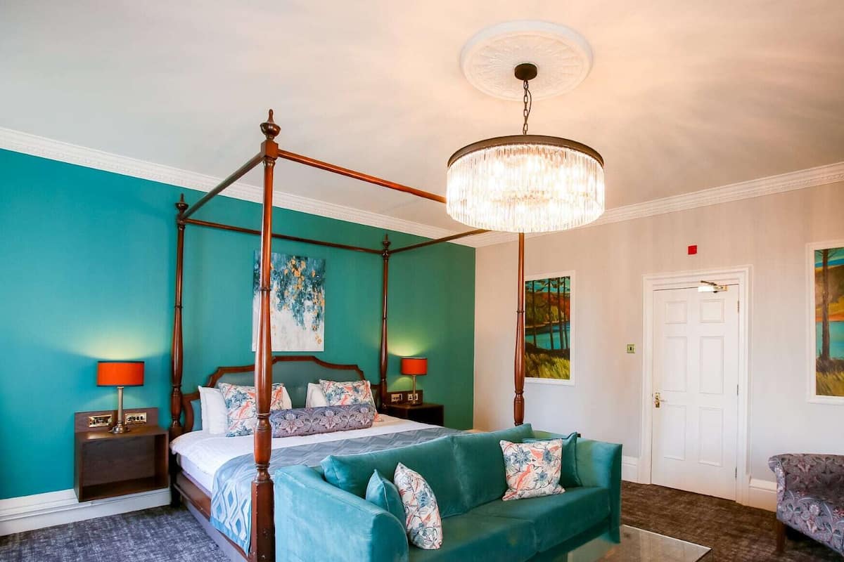 Ein Bett mit Baldachin, eine Kristalllüster, ein türkisfarbenes Sofa, ein gemustertes Sessel und zwei Bilder an den Wänden.