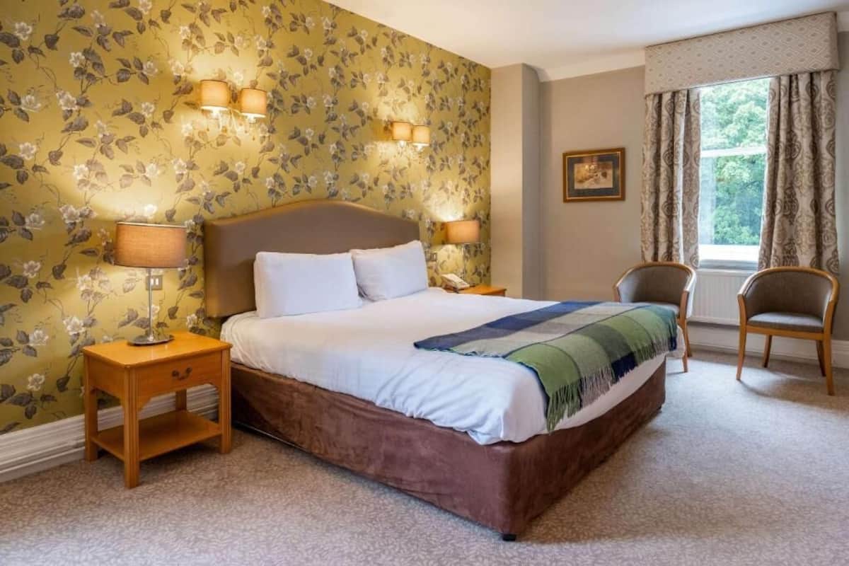 Ein Schlafzimmer mit einem großen Bett, zwei Nachttischen, einem Sessel, blumigem Wandpapier und einem Fenster mit Vorhängen.