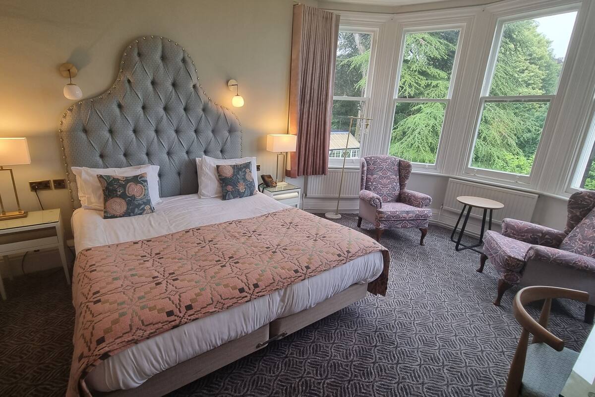 Ein Hotelzimmer mit einem großen Bett, zwei Nachttischen, einem Sessel, einem kleinen Tisch und einem Fenster mit Blick auf Bäume.