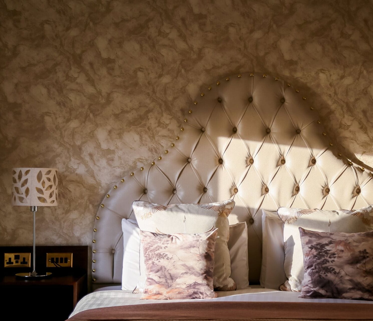 A bedroom with a tufted headboard, multiple pillows, a bedside table with a lamp, and a textured wall.