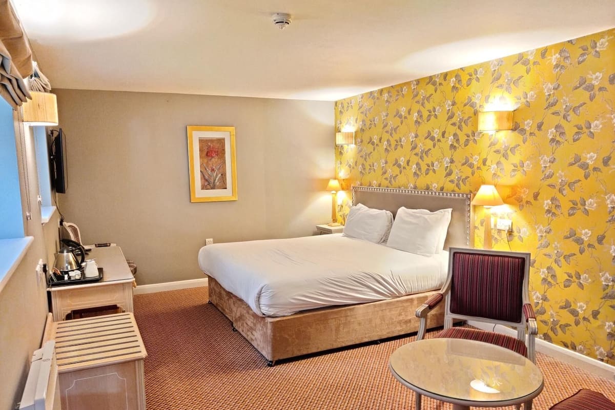 Ein Hotelzimmer mit Bett, Nachttischlampen, einem kleinen Tisch, einem Sessel, einem Schreibtisch mit Lampe und einem an der Wand befestigten Fernseher.