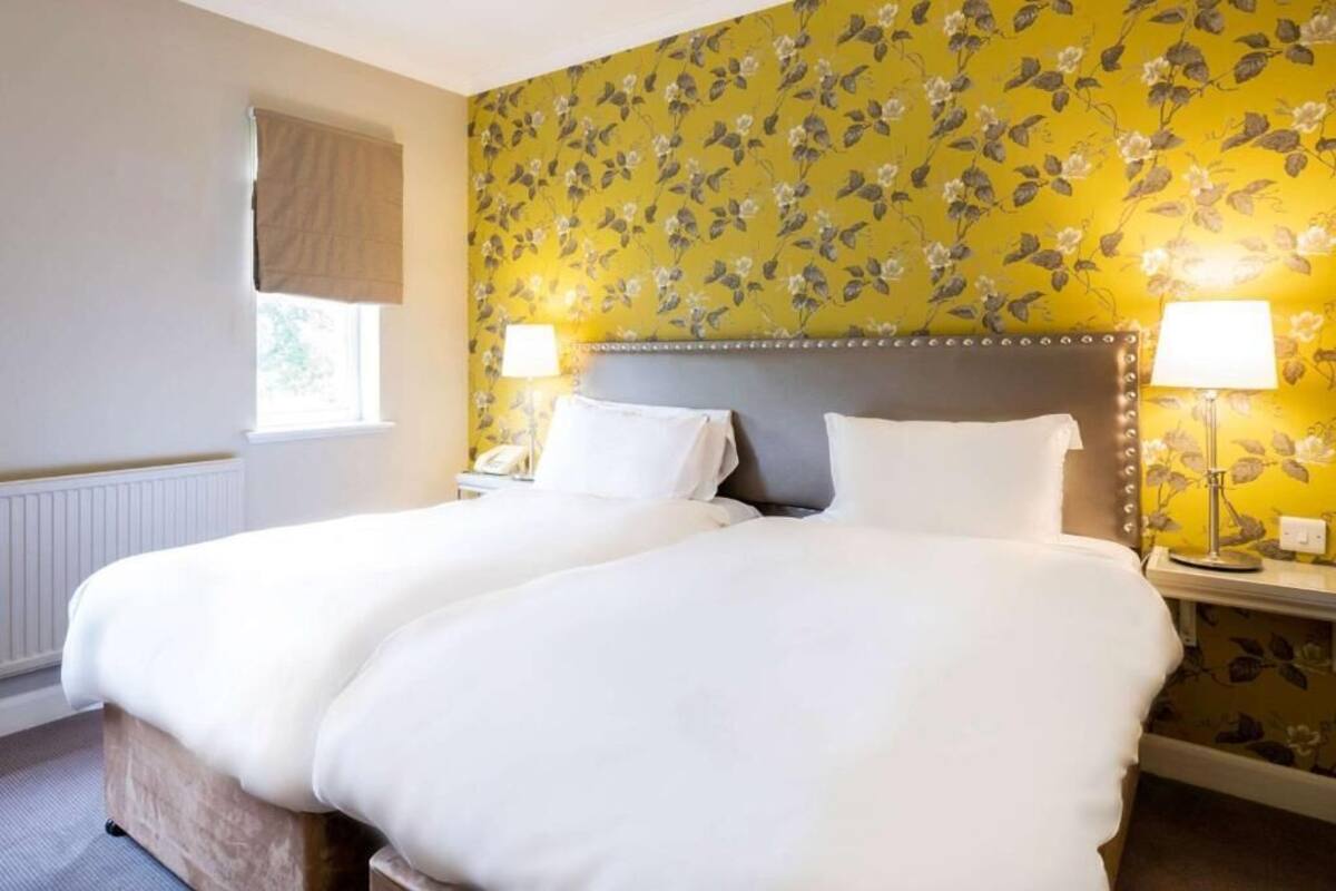 Ein Doppelbett mit weißer Bettwäsche, ein Kopfteil, zwei Nachttischlampen, ein Fenster mit einem beige farbenen Vorhang und floraler Tapete.