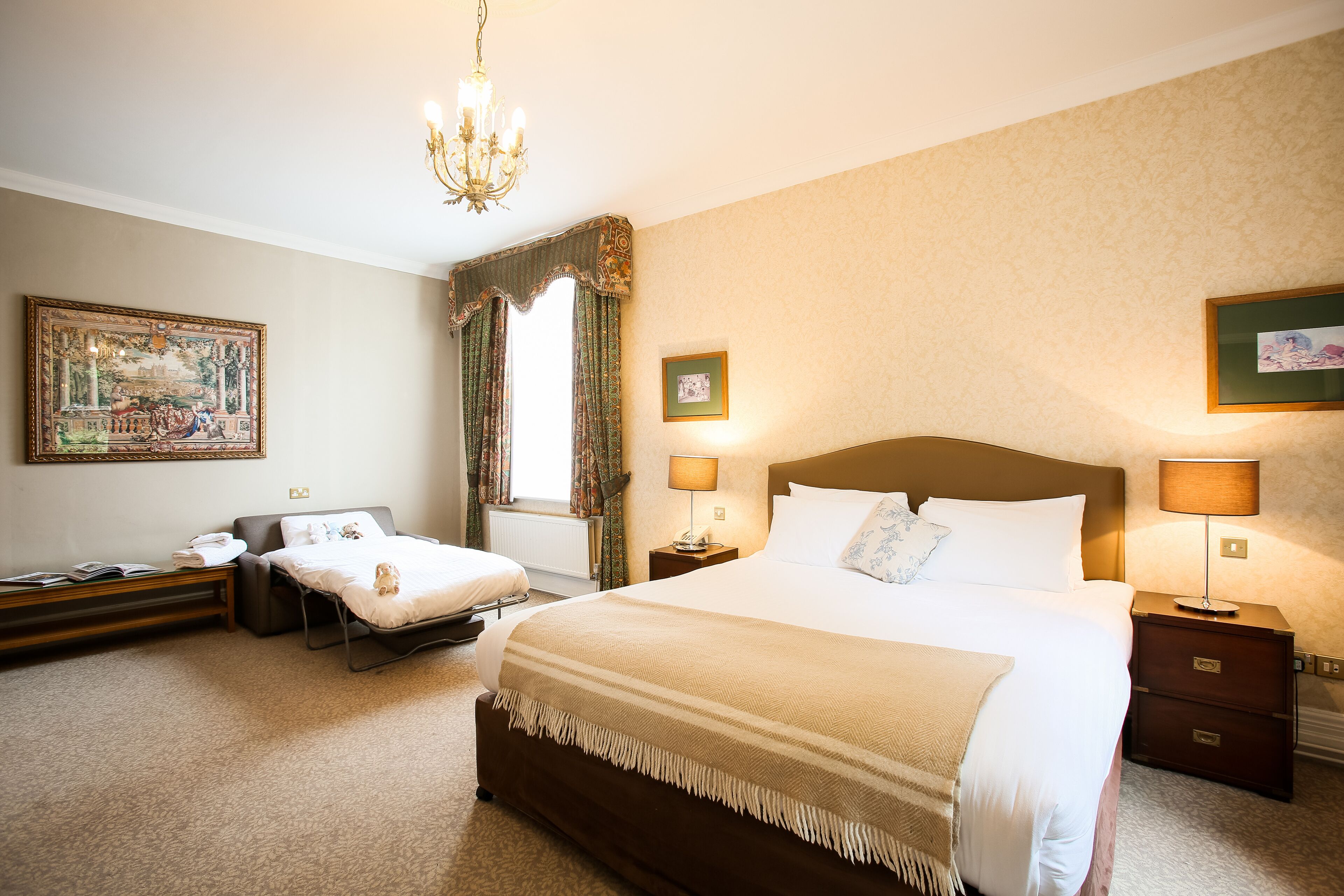 Una habitación de hotel con una cama grande, dos mesitas de noche, un escritorio, una silla, un ventanal con cortinas, un candelabro y obras de arte en las paredes.