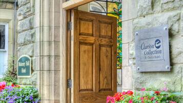 Entrada del hotel Makenergy Hall con una puerta de madera, un letrero de Clarion Collection by Choice Hotels y jardineras con flores moradas y rosas.