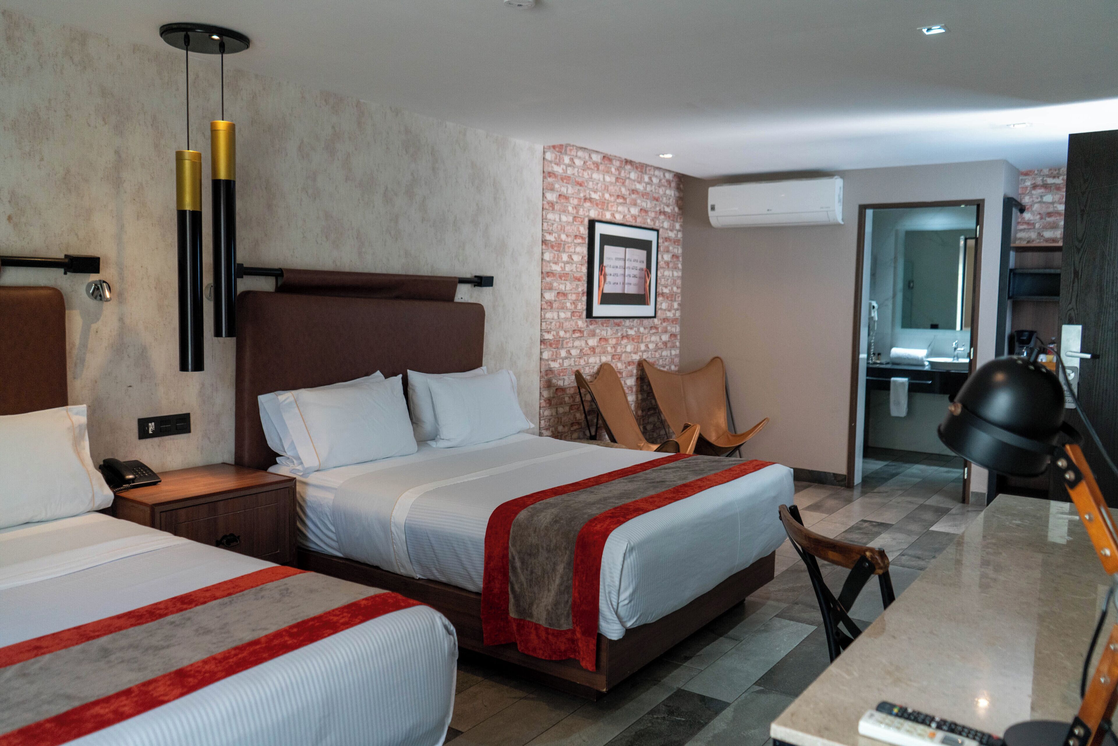 Une chambre d’hôtel avec deux lits, un bureau, une chaise, un miroir et une salle de bain visible à travers une porte ouverte.