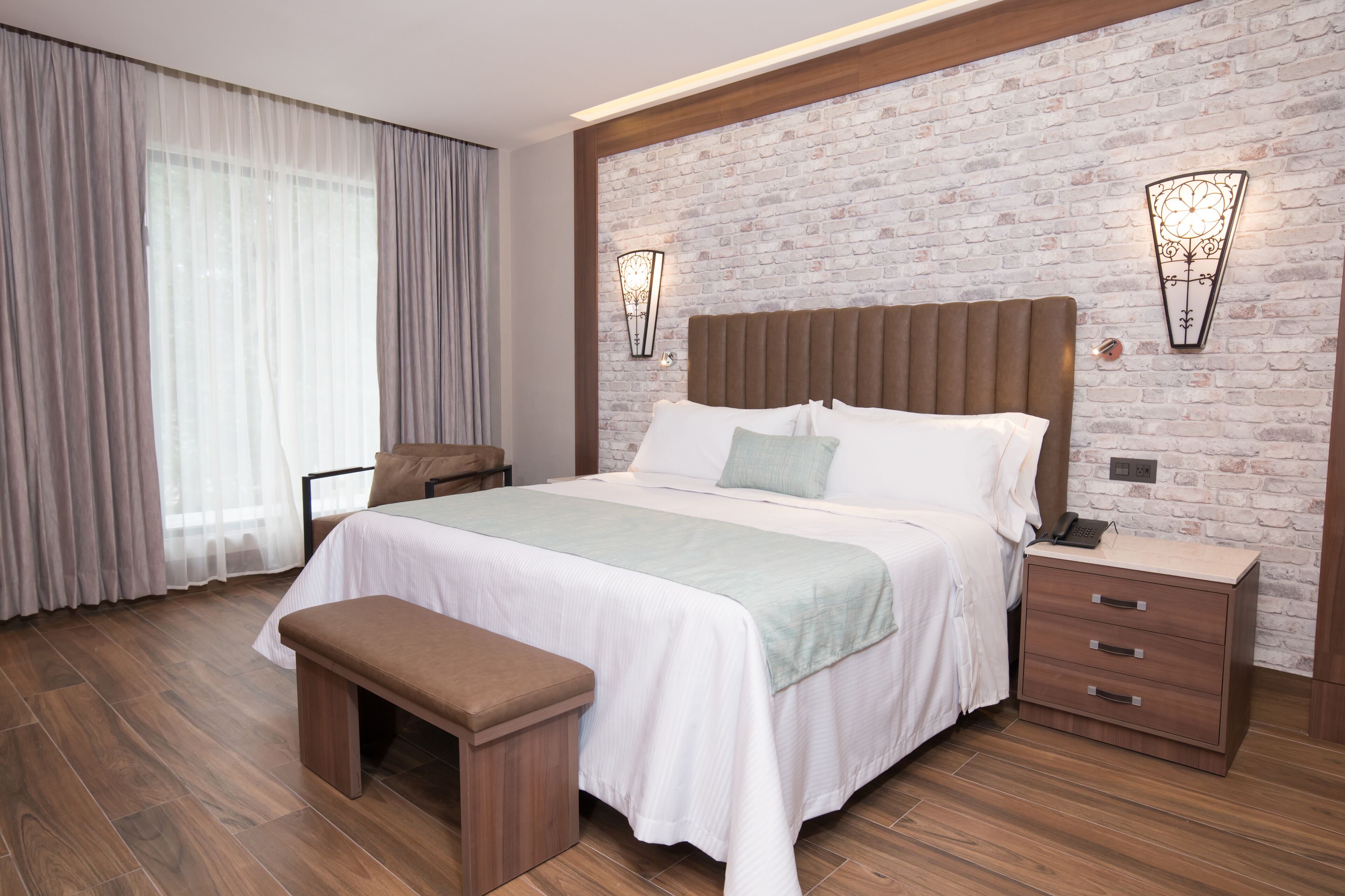 A modern bedroom with a large bed, wooden headboard, two bedside tables, a bench, and a brick-patterned wall.