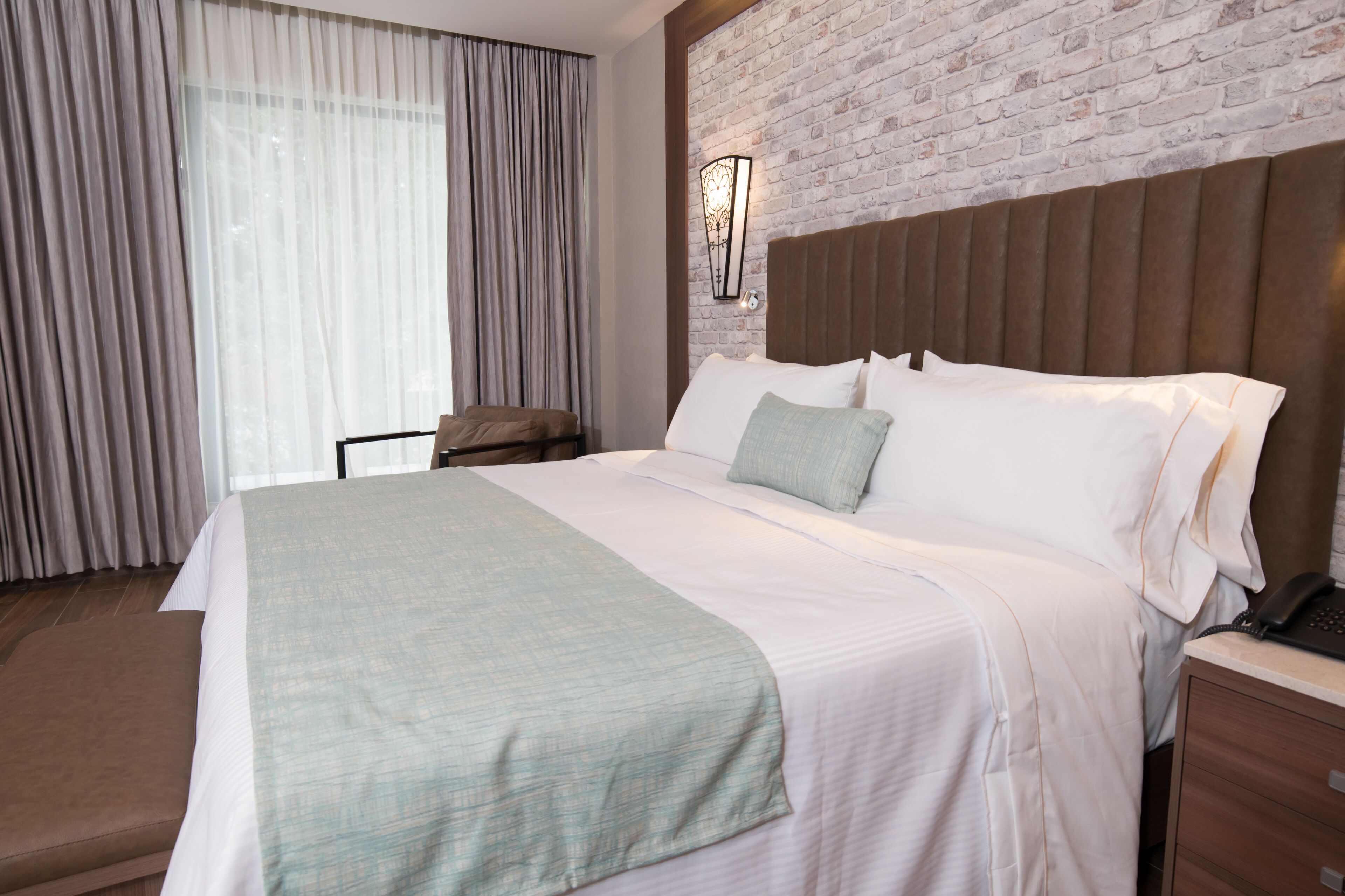 Habitación de hotel con una cama grande, cabecera, mesita de noche, lámpara y una ventana con cortinas.