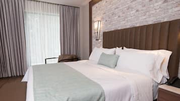 Habitación de hotel con una cama grande, cabecera, mesita de noche, lámpara y una ventana con cortinas.