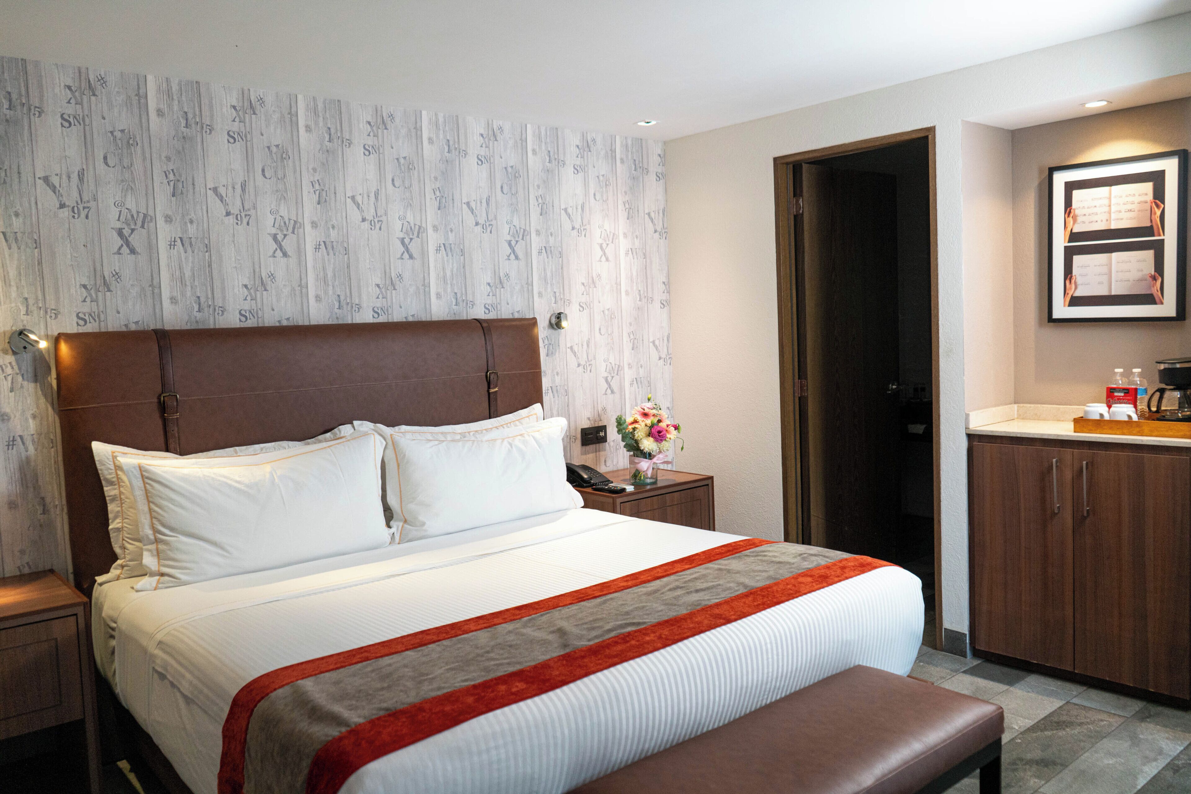 Habitación de hotel con una cama grande, cabecera de madera, mesita de noche con un teléfono y un florero, baño con lavamanos y armarios, y un cuadro enmarcado en la pared.