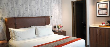 Habitación de hotel con una cama grande, cabecera de madera, mesita de noche con un teléfono y un florero, baño con lavamanos y armarios, y un cuadro enmarcado en la pared.