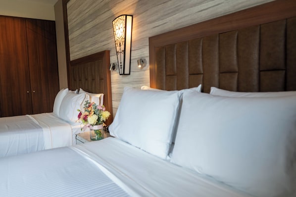 Habitación de hotel con dos camas, un cabecero de madera, una lámpara de pared y una mesita de noche con un florero.