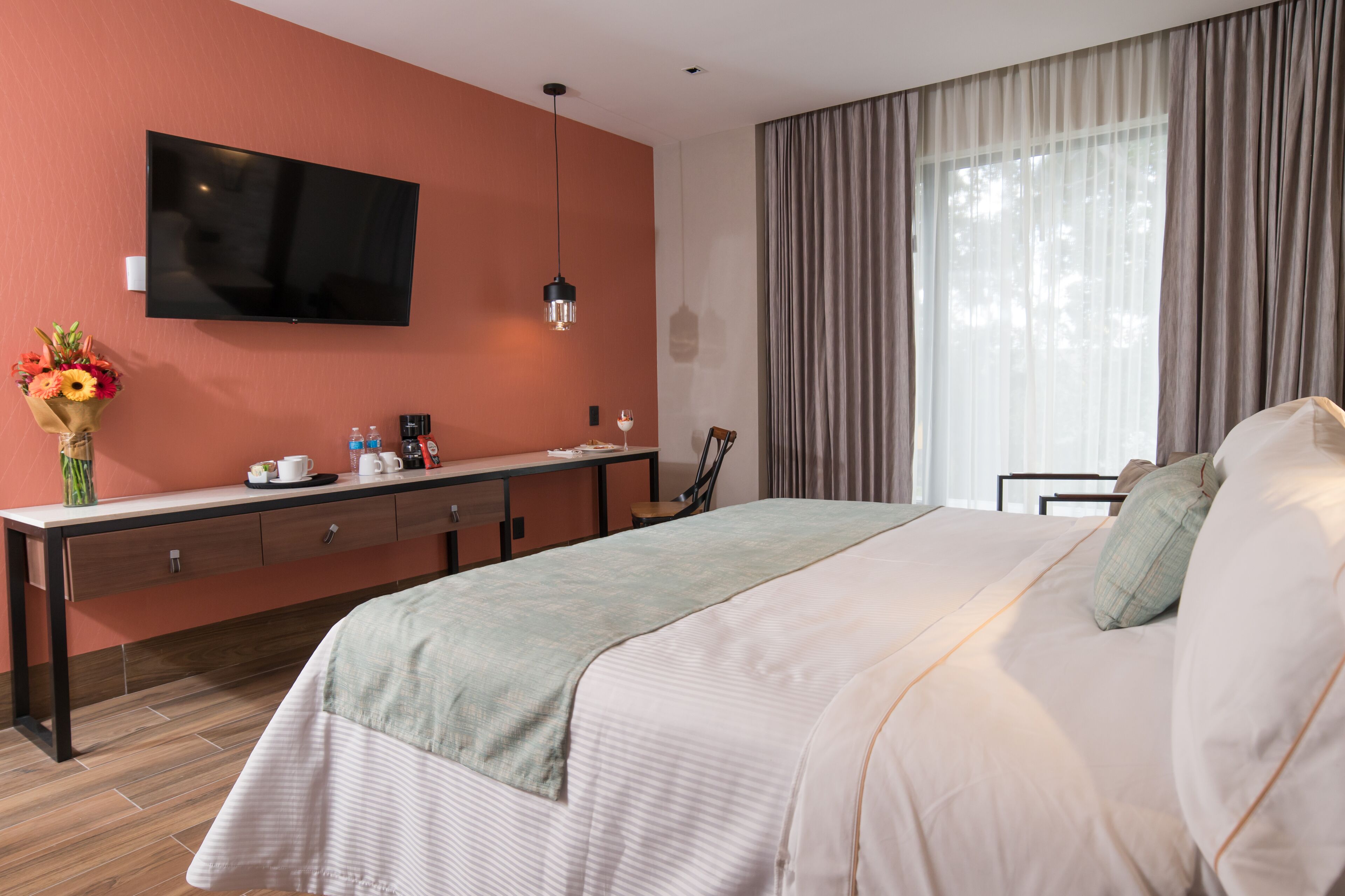 A hotel room with a large bed, a desk with a flat-screen TV, a vase with flowers, and a window with curtains.