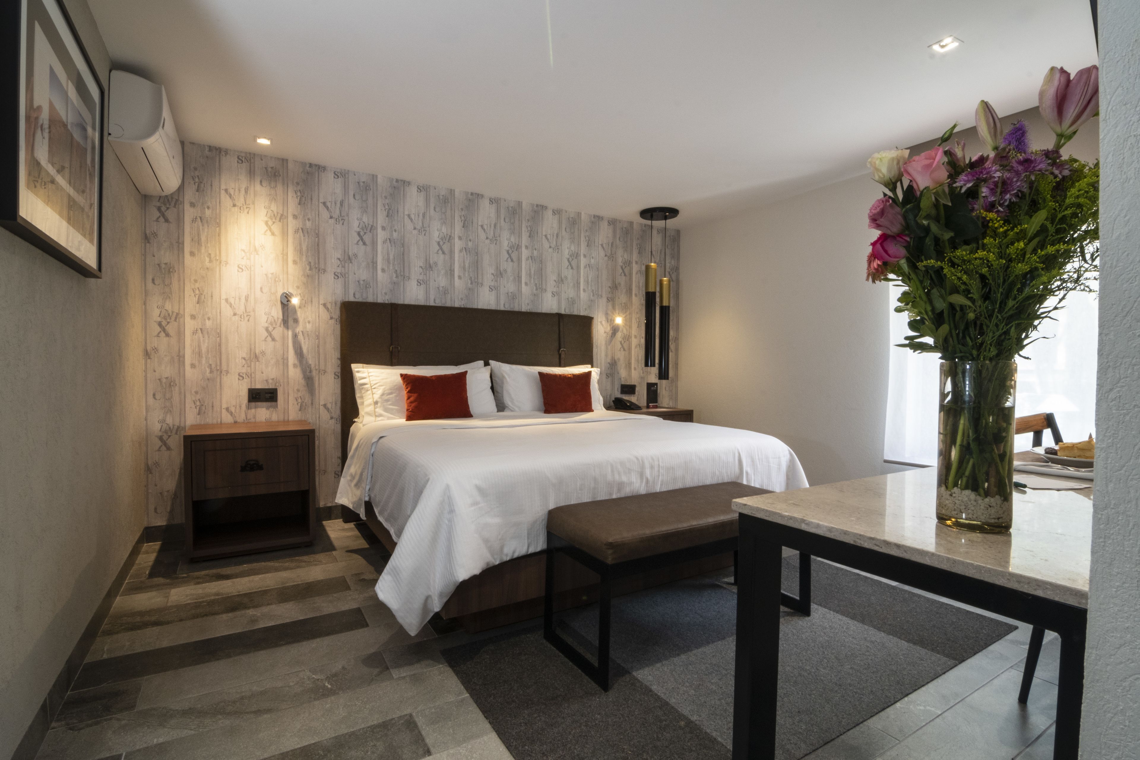 Une chambre d’hôtel moderne avec un grand lit, des tables de chevet, un banc, un vase avec des fleurs et un tableau encadré au mur.