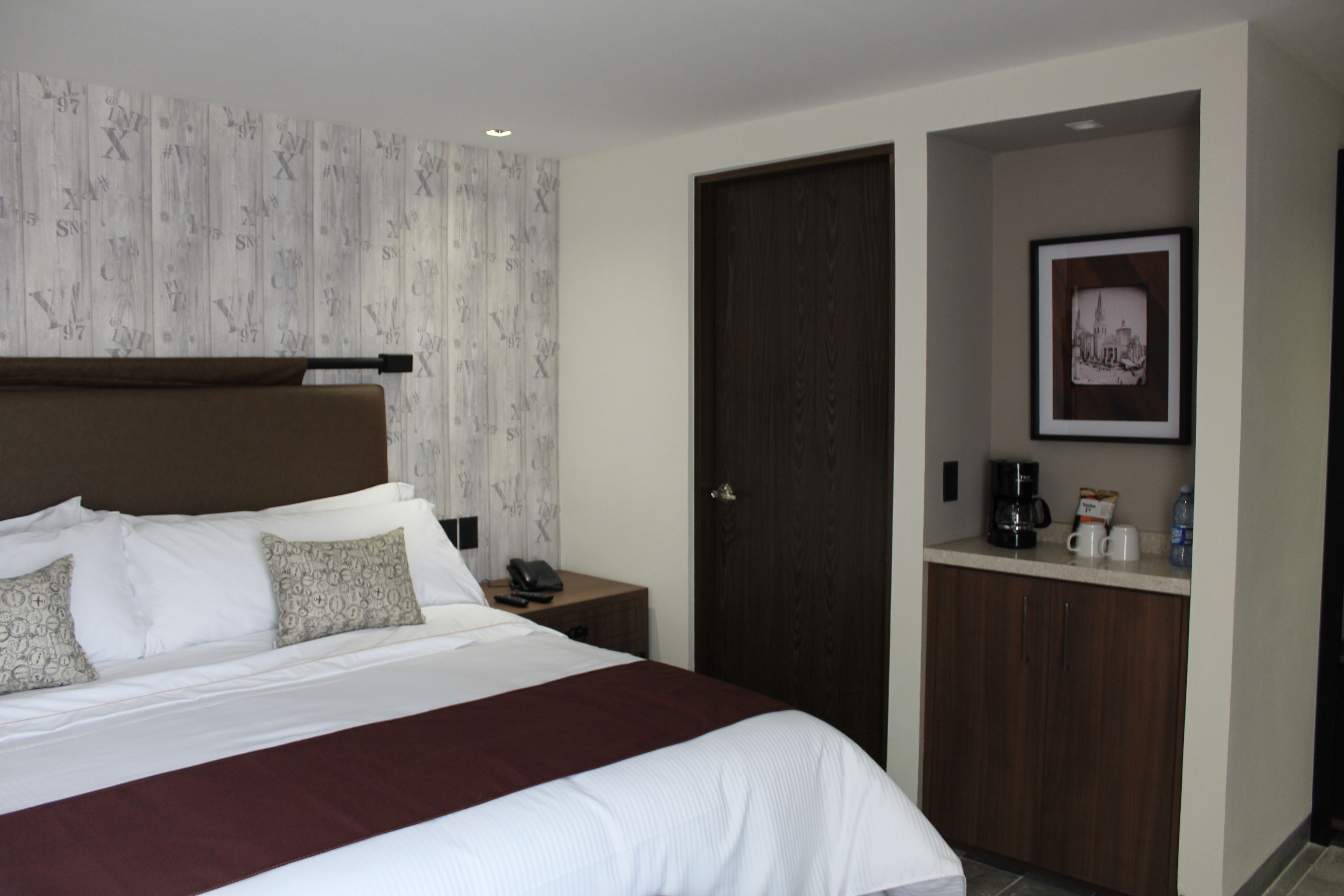 Habitación de hotel con una cama grande, cabecera de madera, mesita de noche con teléfono y cafetera, un cuadro en la pared y una puerta a la derecha.