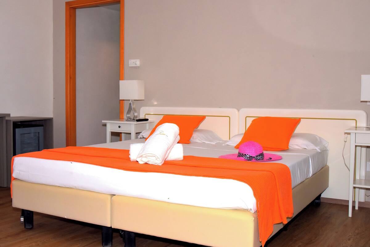 Cama doble con colcha y almohadas anaranjadas, mesita de noche blanca con lámpara y un escritorio pequeño con una computadora.