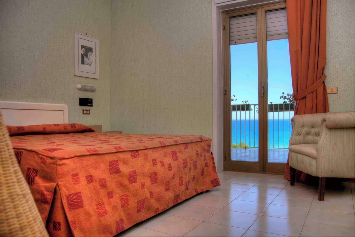 Habitación de hotel con cama, una silla, un balcón y vistas al mar, además de una puerta.