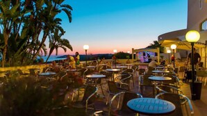 An outdoor patio area with round tables and chairs, palm trees, and a sunset view. - Paradise Beach Resort (Castelvetrano)