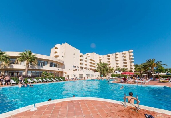 A swimming pool with lounge chairs and palm trees, surrounded by a multi-story hotel building.