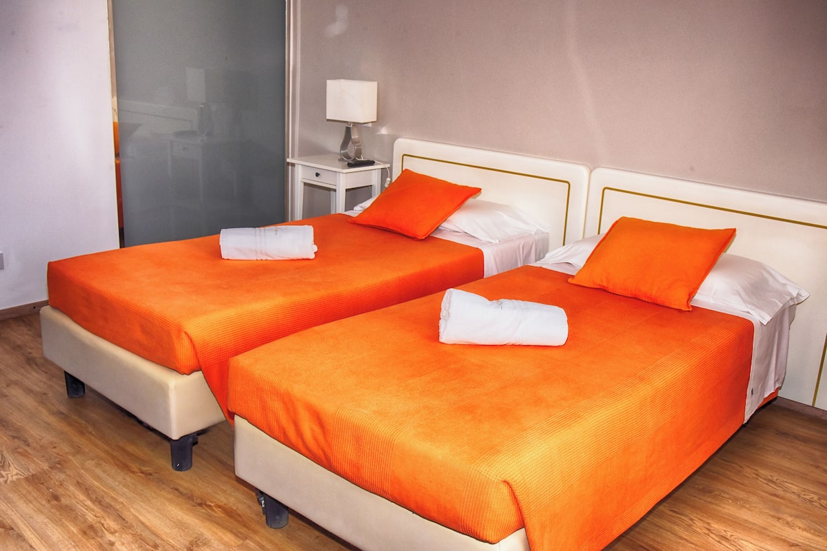 Dos camas con ropa de cama naranja y almohadas blancas, una mesita de noche pequeña con una lámpara, y piso de madera.