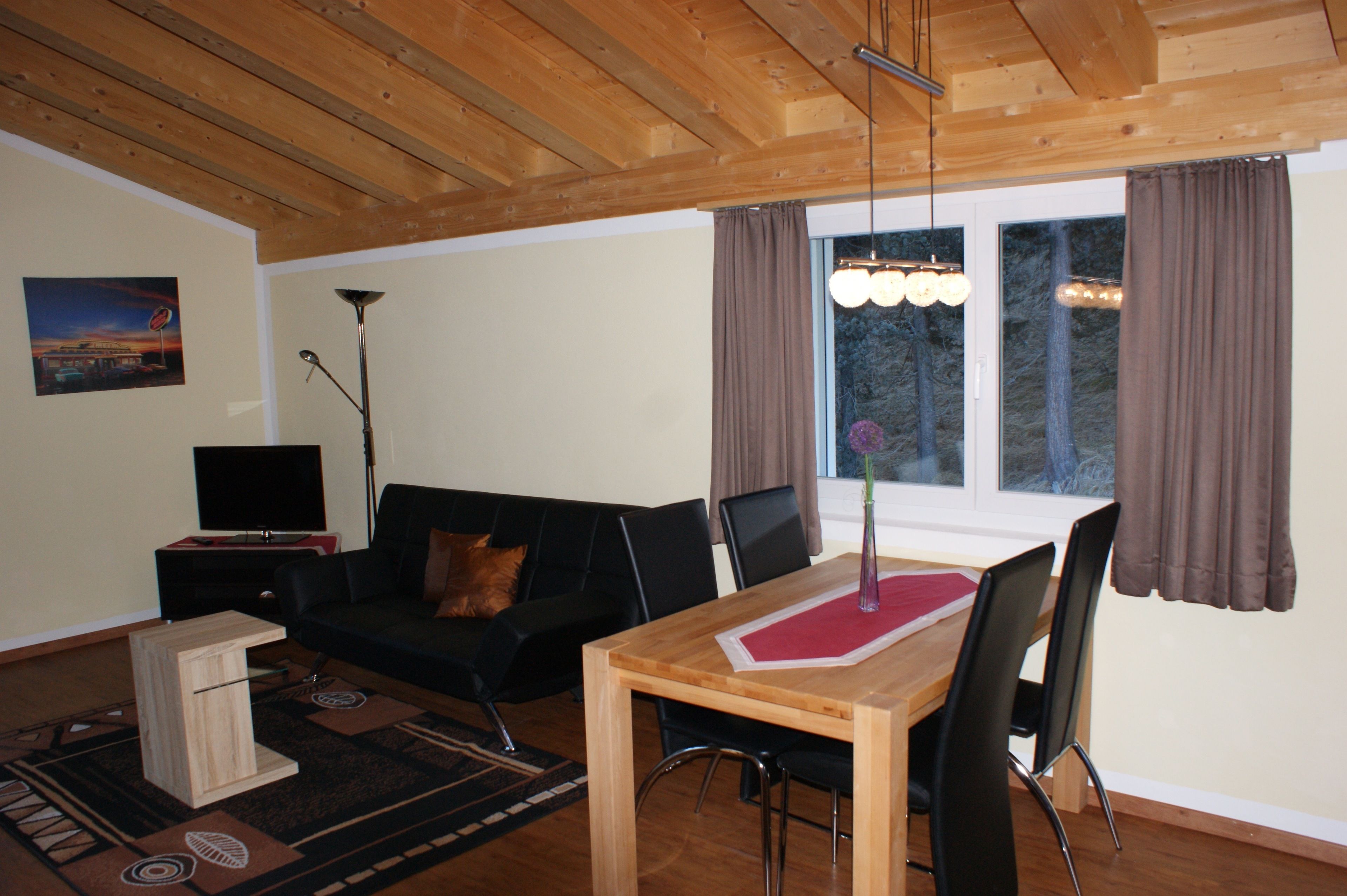 A living room with a wooden ceiling, a black sofa, a wooden dining table with chairs, a TV on a stand, and a window with curtains.