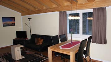 A living room with a wooden ceiling, a black sofa, a wooden dining table with chairs, a TV on a stand, and a window with curtains.