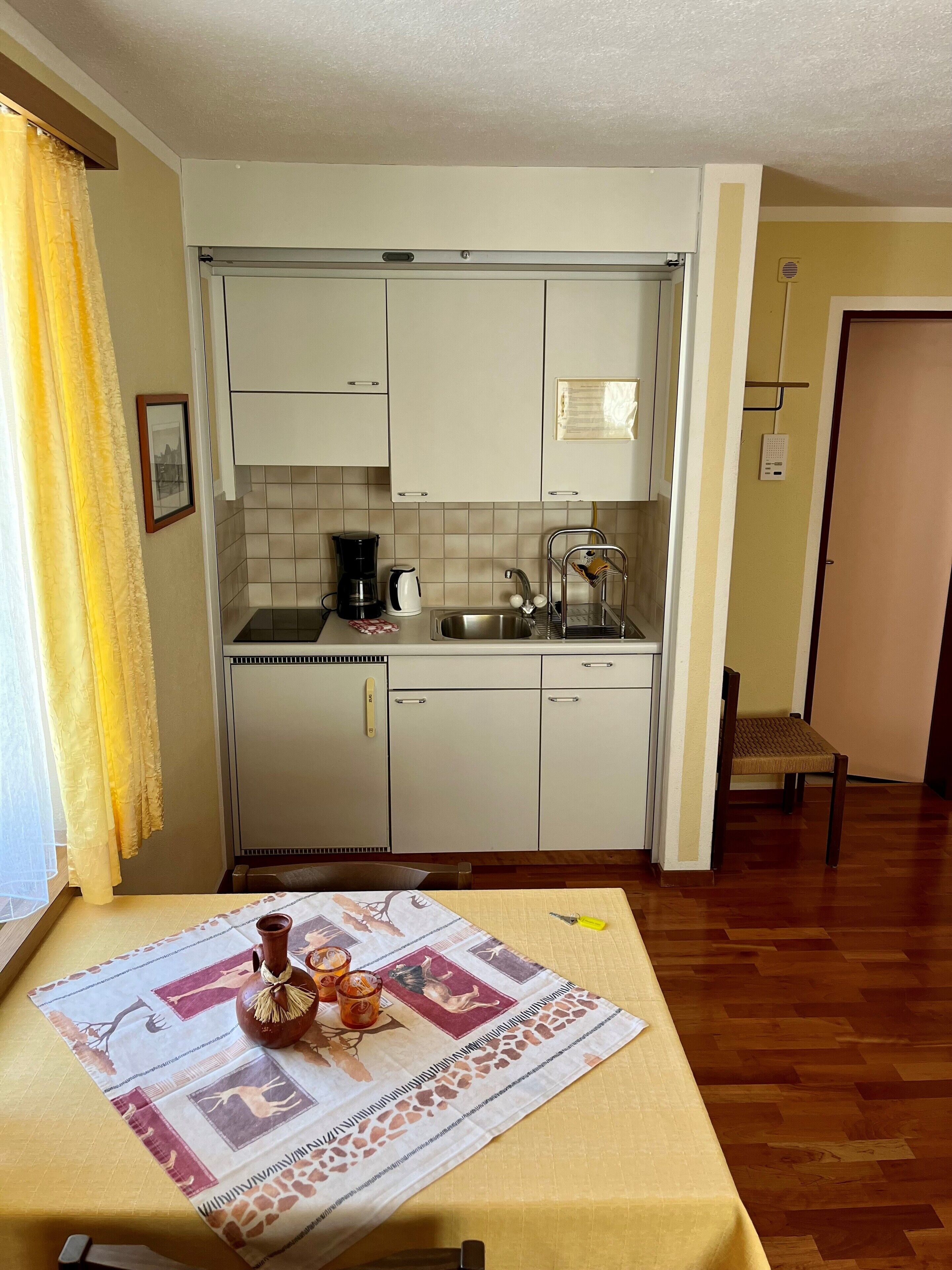 A compact kitchen with white cabinets, a sink, and a coffee maker. A dining table with a placemat and a vase is in the foreground.
