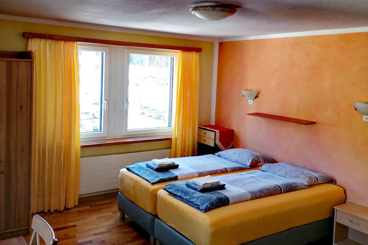 A room with two beds, wooden flooring, a window with curtains, a small table, and a wall-mounted shelf.