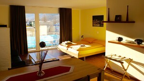 Una habitación con cama, escritorio, silla, ventana con cortinas, una mesita con un florero y estante con adornos.