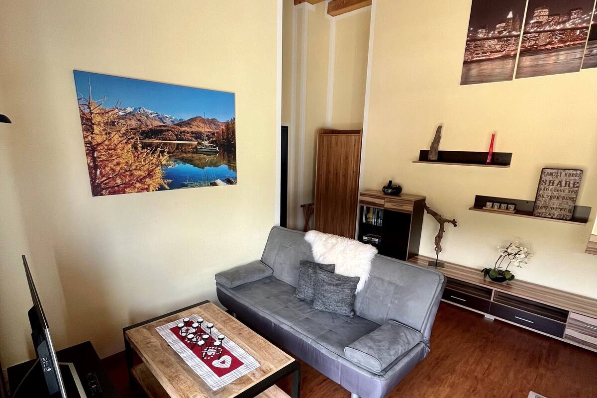 A living room with a grey sofa, wooden coffee table, and a TV stand. There are wall-mounted shelves with decorative items and a large landscape painting.