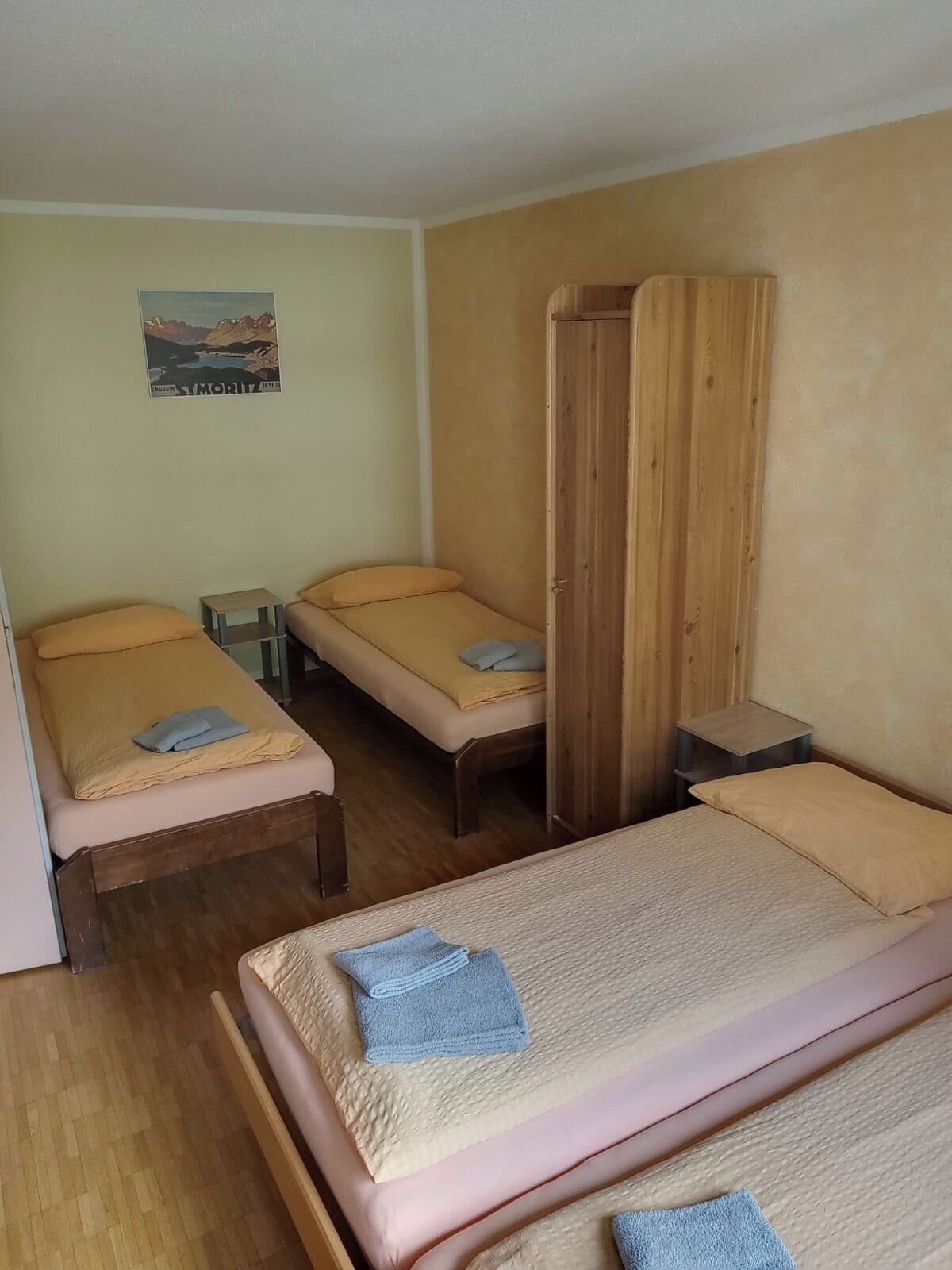 A small, simple room with three single beds, wooden furniture, and a poster on the wall.