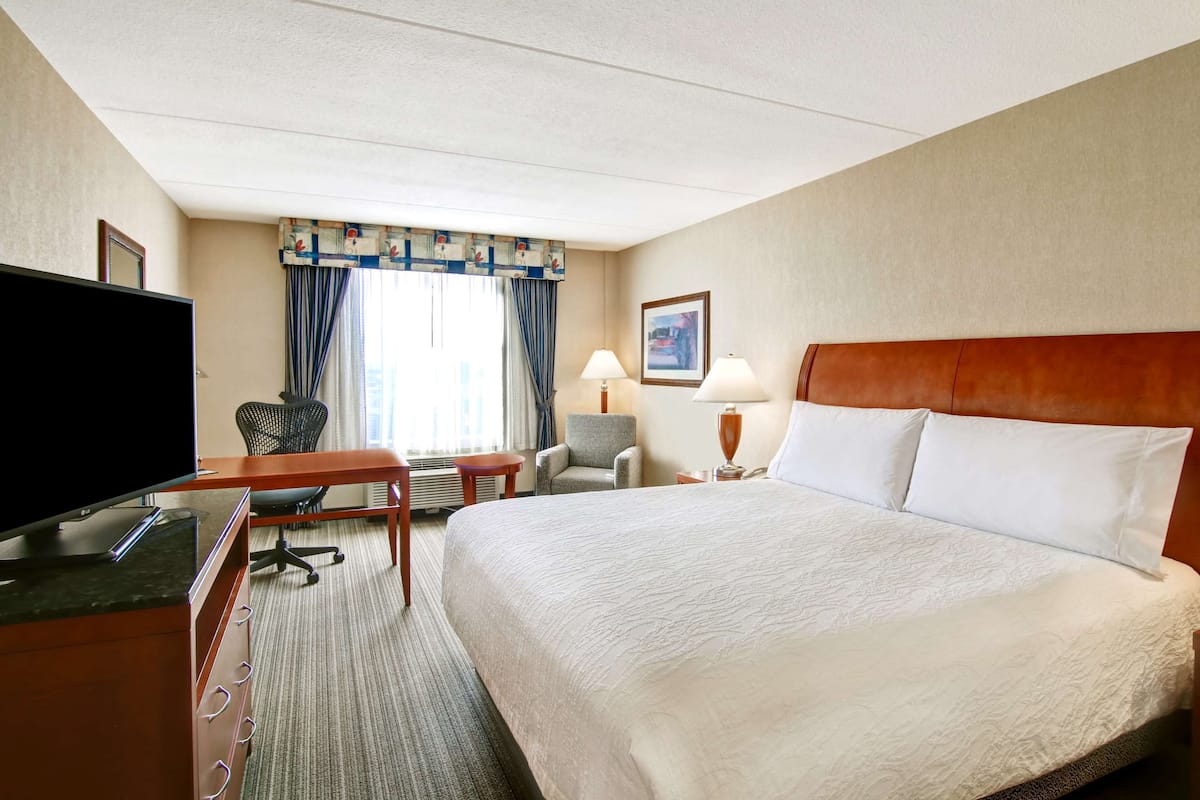 A hotel room with a large bed, a desk with a flat-screen TV, two chairs, a window with curtains, and a painting on the wall.