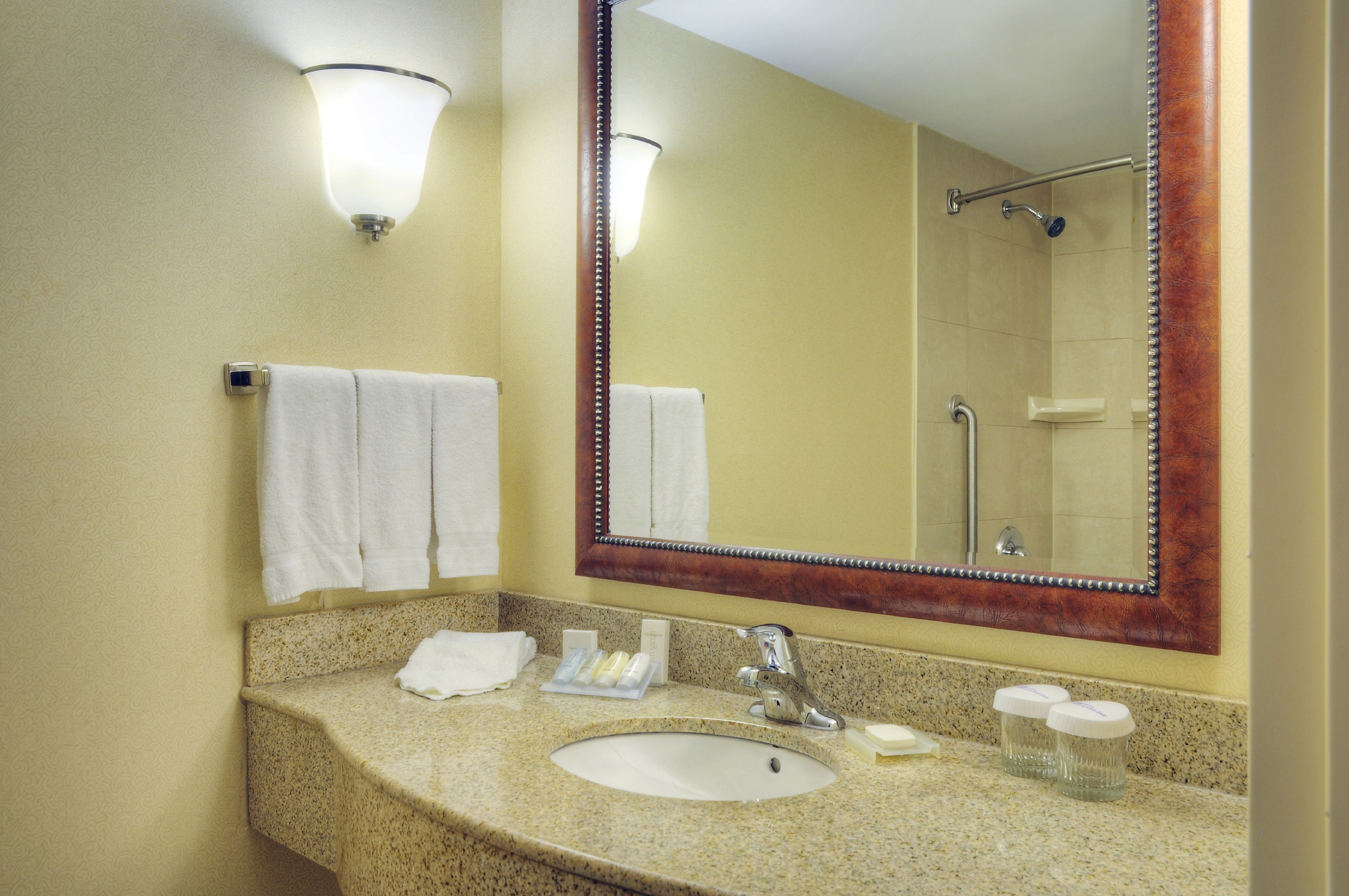 Une salle de bain dotée d’un comptoir en granit, d’un lavabo blanc, d’un grand miroir avec un cadre en bois et de deux appliques murales.