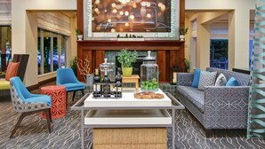 A lobby with a large TV screen, seating area with sofas and chairs, a coffee table with bottles, and a fireplace. - Hilton Garden Inn Toronto/Burlington (Burlington)
