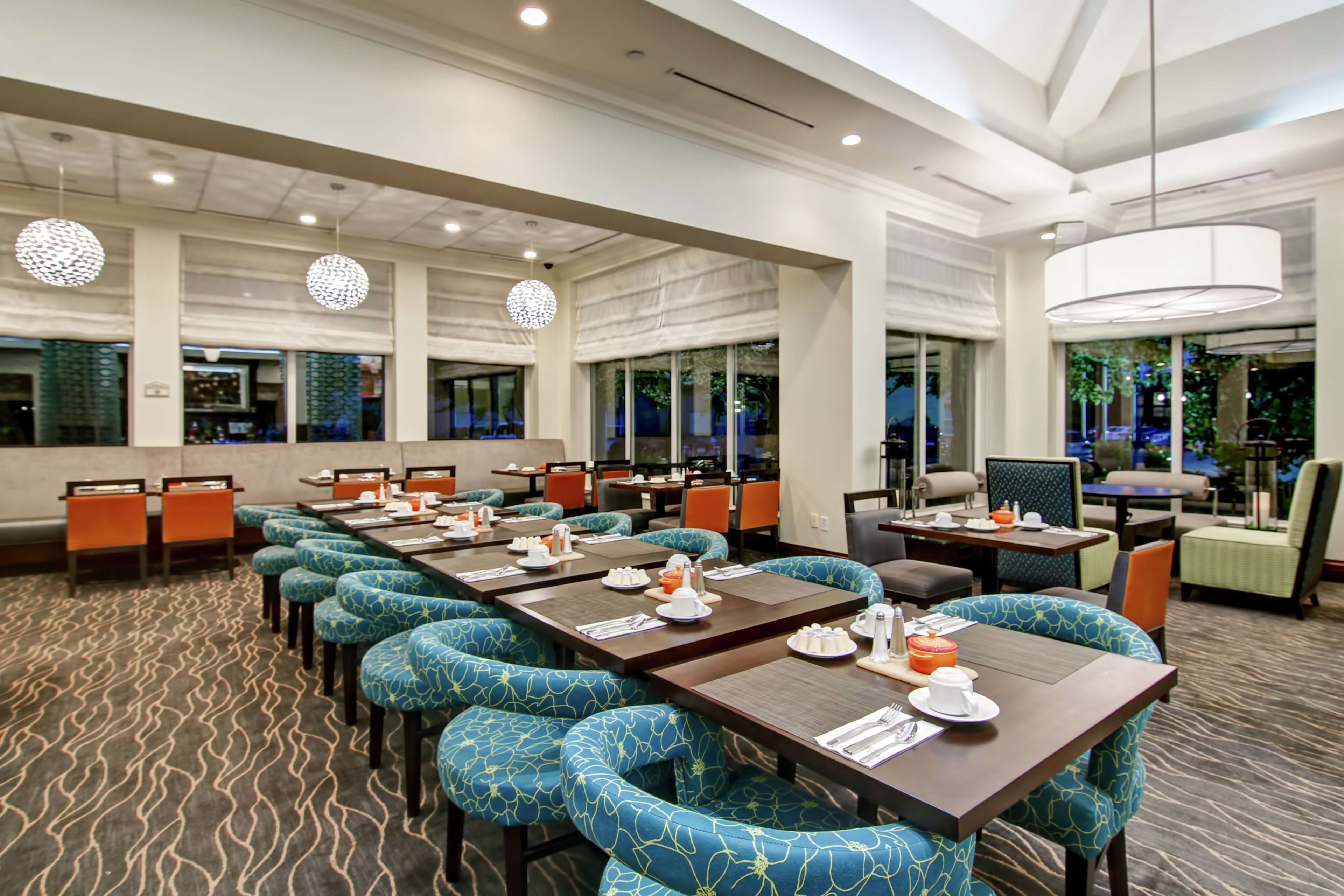 Um espaço de refeições de hotel com várias mesas e cadeiras, grandes janelas e um tapete estampado.