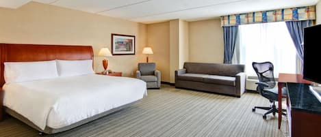 A hotel room with a large bed, a sofa, a desk with a chair, a TV, and a window with curtains.