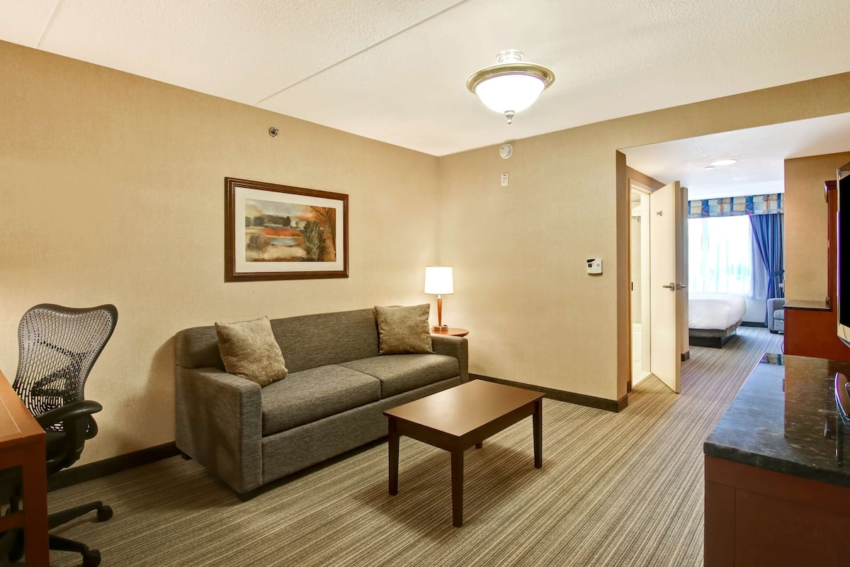 A hotel room with a sofa, a wooden coffee table, a framed picture on the wall, a lamp, and a view of the adjacent bedroom.