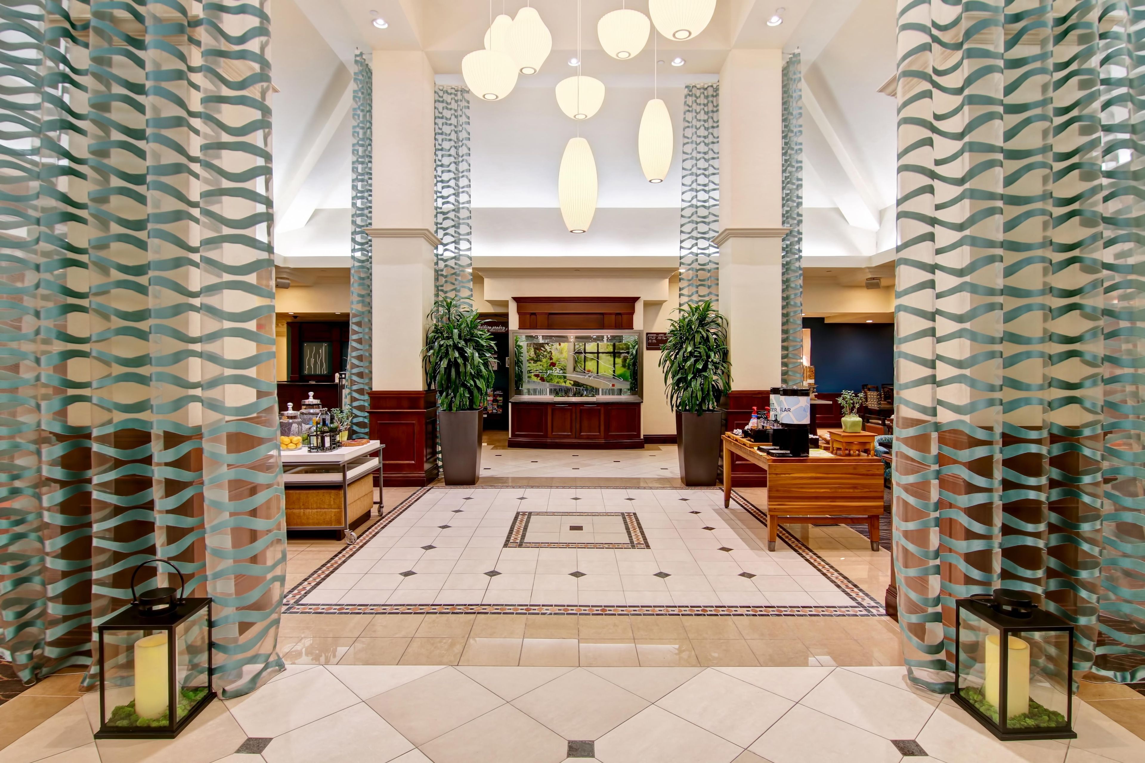 A spacious lobby with a central reception desk, potted plants, and a tiled floor.