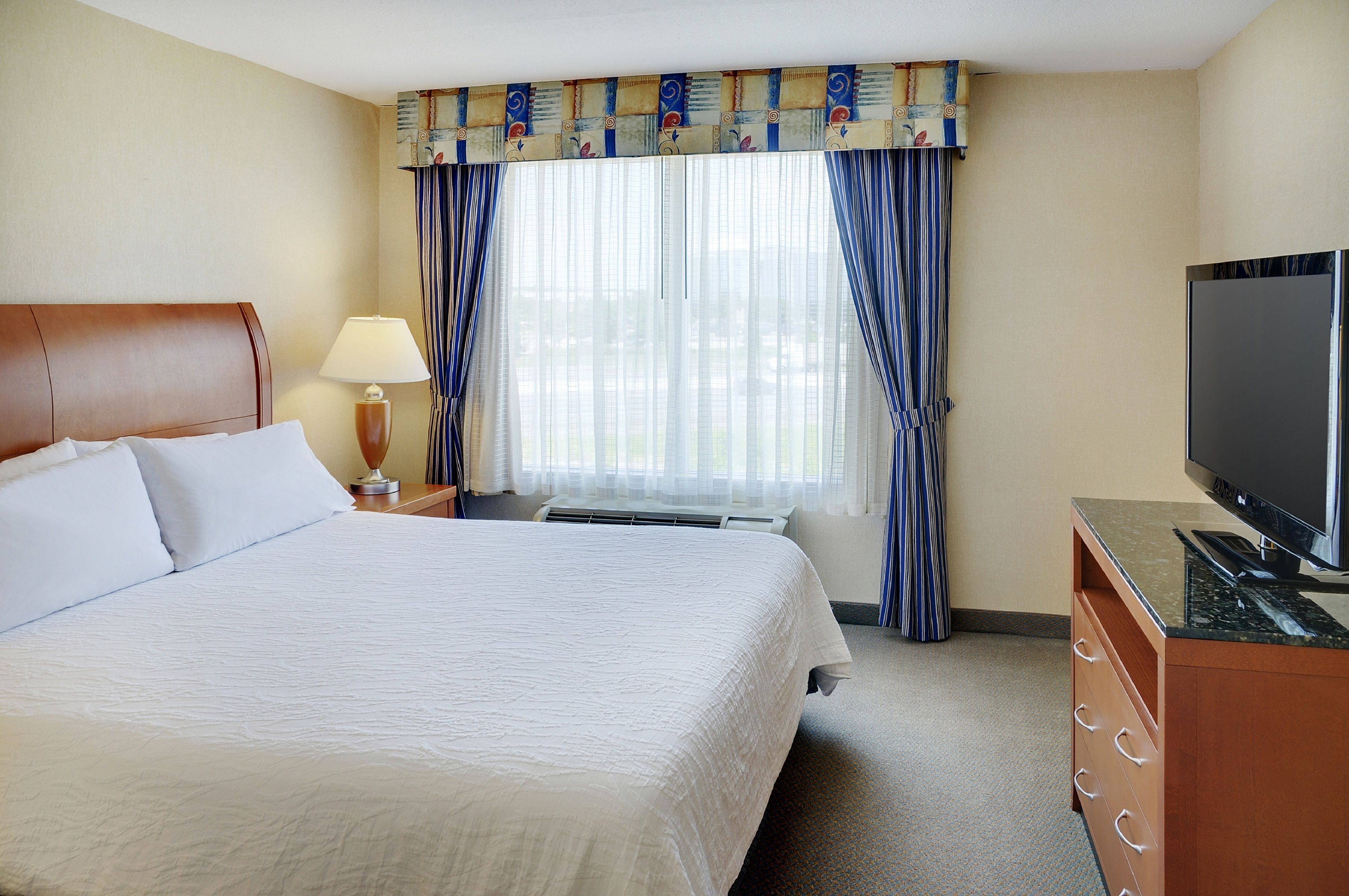 A hotel room with a large bed, a flat-screen TV, a desk with a lamp, and a window with curtains.