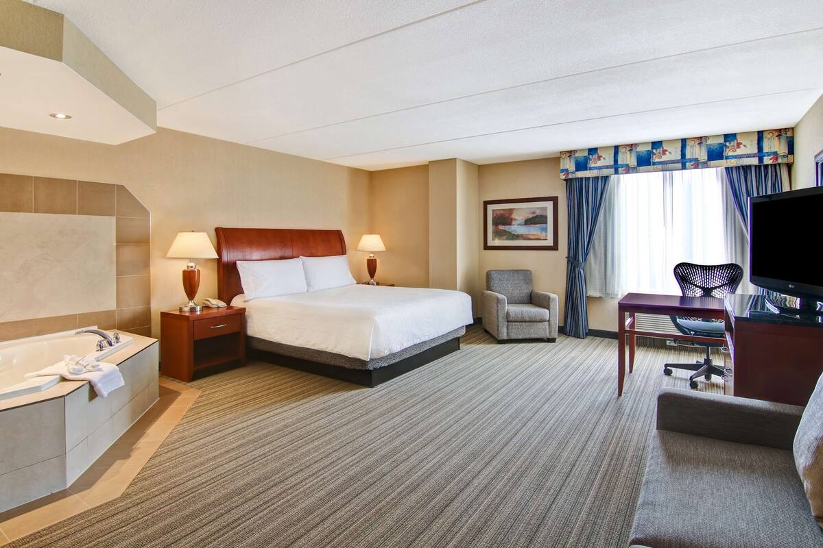 A hotel room with a large bed, a desk with a computer, a chair, a TV, a bathroom with a bathtub, and a window with curtains.