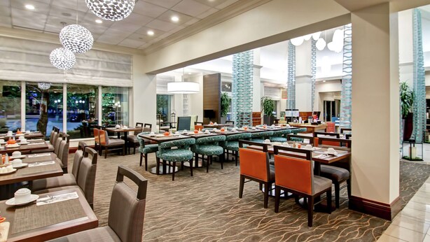 A spacious dining area with multiple tables and chairs, large windows, and decorative lighting fixtures. - Hilton Garden Inn Toronto/Burlington (Burlington)