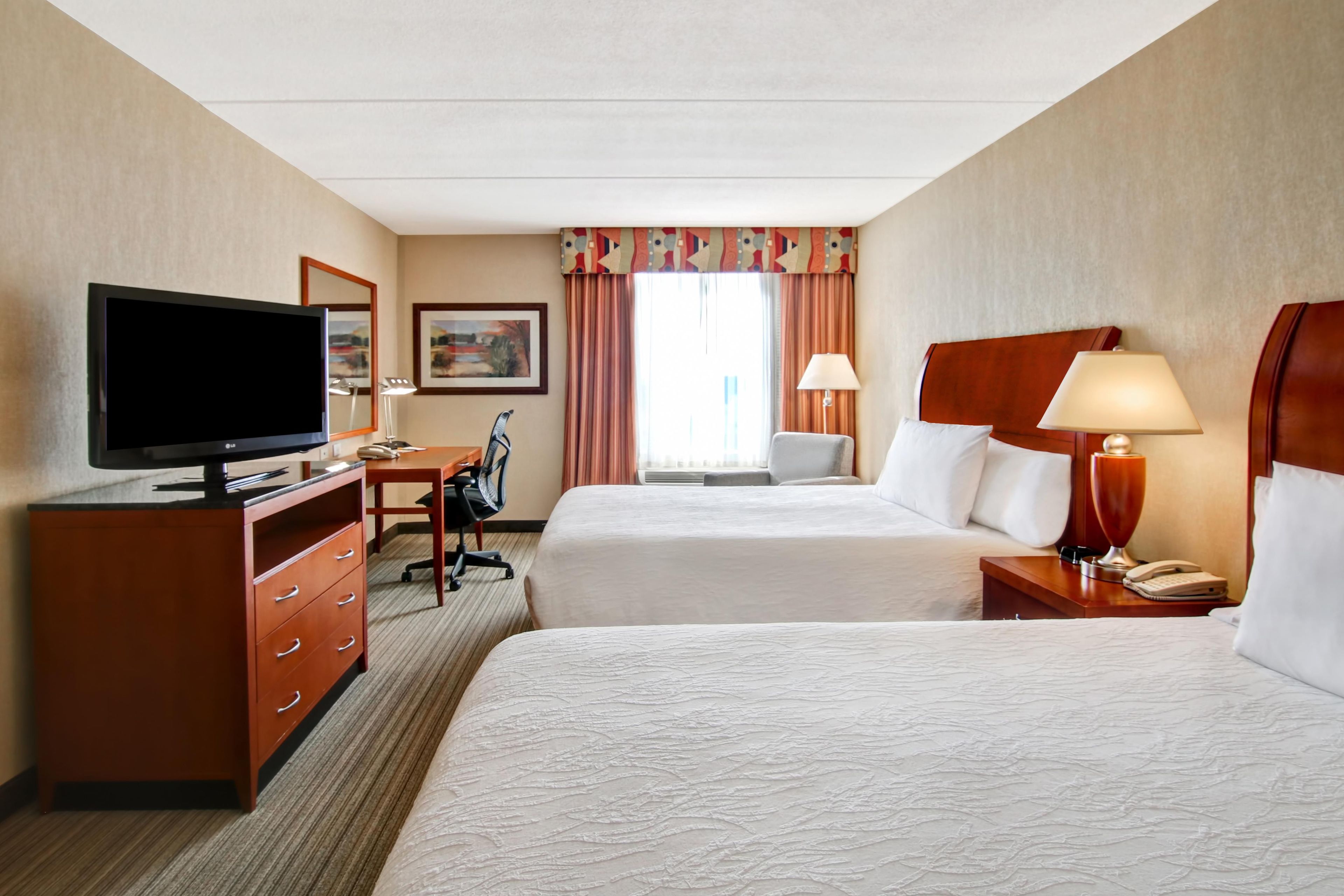 A hotel room with two beds, a desk with a television, a chair, and a lamp.