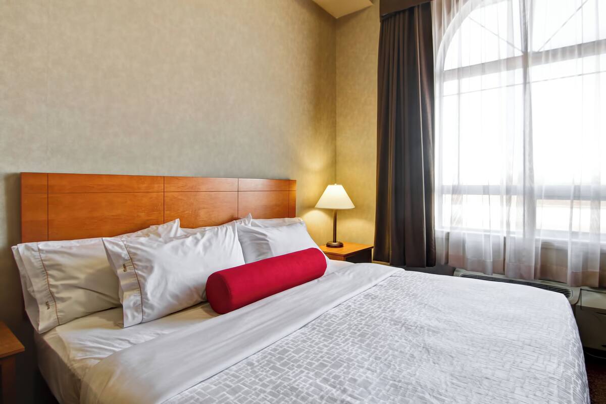 Una cama bien hecha con sábanas blancas y un cojín rojo decorativo, cabecera de madera, lámpara de noche y un ventanal con cortinas de encaje.