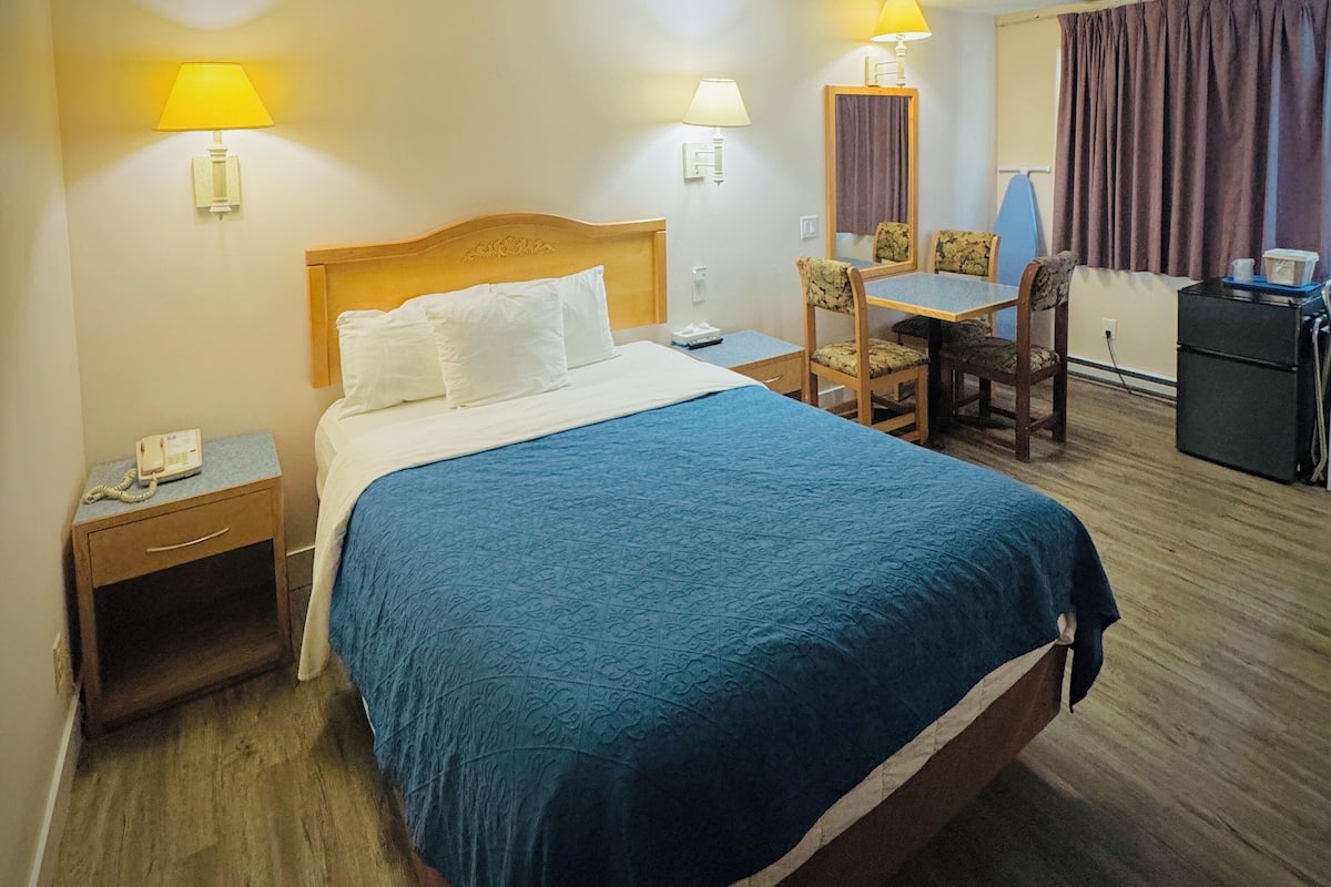 A hotel room with a bed, bedside table, lamp, chair, desk, and a small kitchenette.