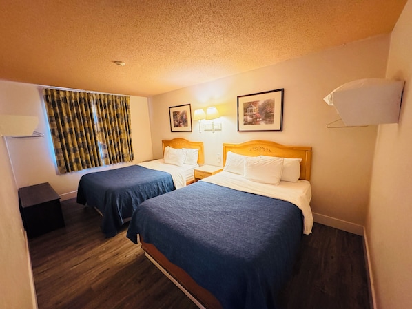 Habitación de hotel con dos camas individuales, cabeceros de madera, una mesita de noche pequeña, una lámpara, un cuadro enmarcado en la pared y una ventana con cortinas.