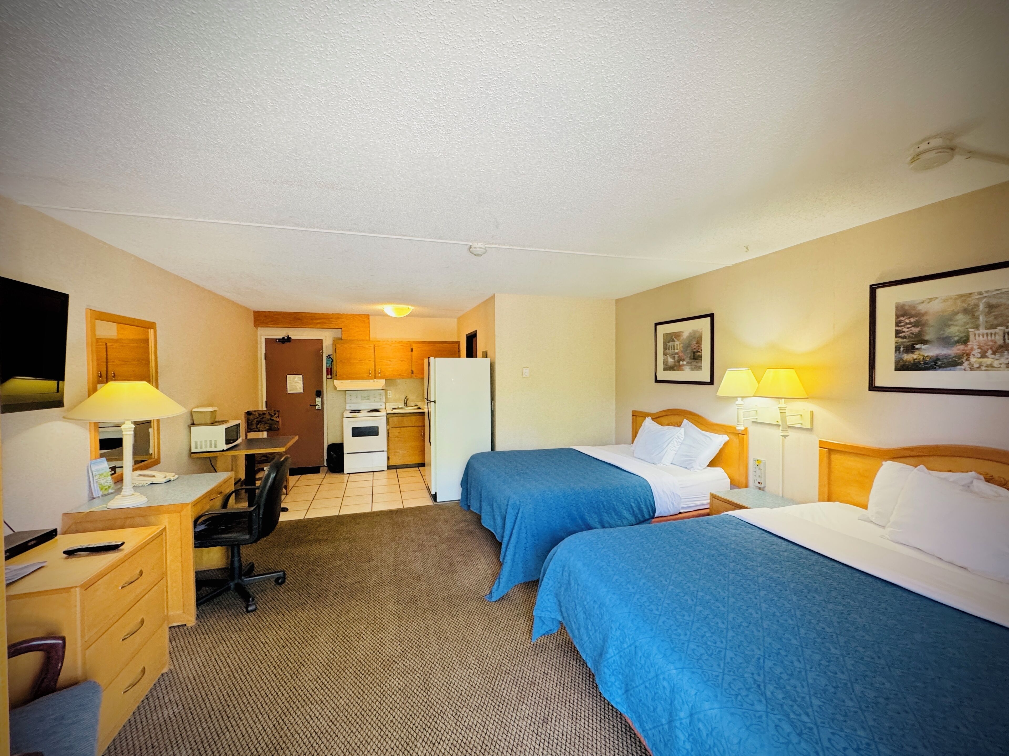 A hotel room with two beds, a desk, a chair, a microwave, a refrigerator, and a small kitchenette.